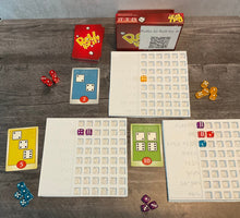 Load image into Gallery viewer, The game is shown setup(red version). There are trays next to the cards to sort who's dice are who's. The cards all have transparent braille on them indicated the needed dice and their point value