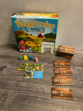 Load image into Gallery viewer, The game box, with the meeples in front of it. One 3d castle is setup and the dominos are labeled on both sides in transparent braille