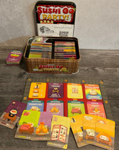Load image into Gallery viewer, The sushi go party game and board. Cards are laid out in front of the board. Both the board and the cards have transparent braille on them
