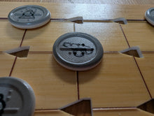 Load image into Gallery viewer, Close-up of round gray tokens placed on the squares. The central token has a cannon symbol printed on it with the letters ca in braille on the piece. There is a full border on the outside of the piece as opposed to the dotted border on the red pieces. Other similar tokens are partially visible around the board.
